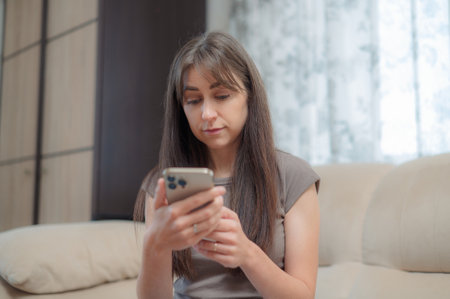 Young woman sitting comfortably in a chair, smiling while using her phone indoors during a relaxing afternoonの写真素材