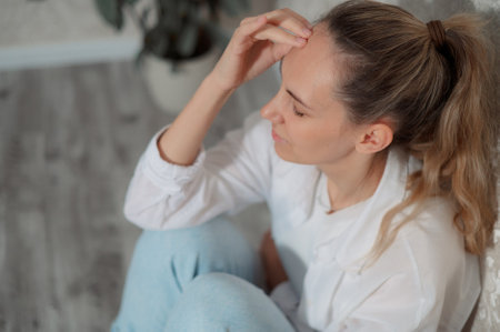 Young woman experiencing headache while sitting on a couch in a modern living room home during daytime vertical photoの写真素材