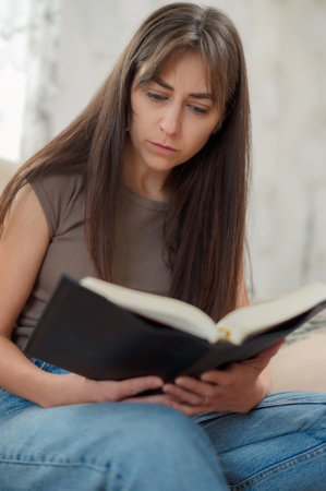Young woman engrossed in a captivating book, sitting comfortably on a sofa in the cozy atmosphere of her living roomの写真素材