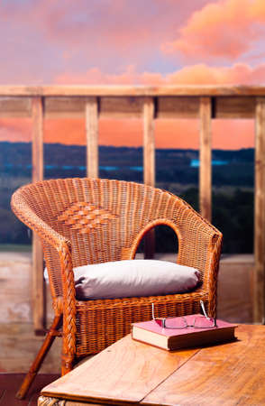 Wicker chair with pillow next to a wooden table with a book and reading glasses on it, with a view of a river at sunset in the backgroundの写真素材