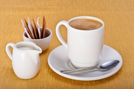 Mug of coffee with saucer, teaspoon, milk jug and sugar sachets on a golden table cloth の写真素材