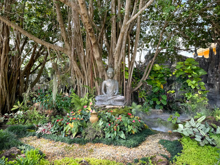 Sacred holy Buddha statue sitting underneath Bodhi tree or Banyan Tree, a tree that is sacred and hallowed for Buddhism teaching, in outdoor park.の写真素材