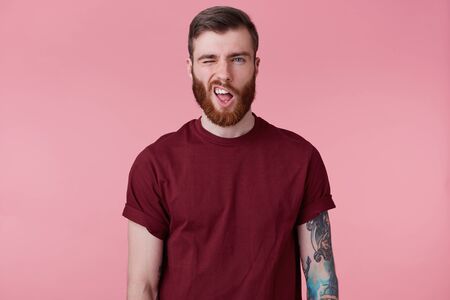 Portrait of young bearded male with tattooed hand makes a frown, screwing eye, isolated over pink background.の写真素材