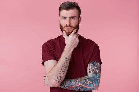 Close up of handsome thoughtful bearded guy with tattooed hand, looking at camera, holding his chin,...