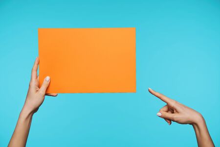 Young attractive hand with white manicure holding piece of paper with raised hand and showing on it with index finger, isolated over blue backgroundの写真素材