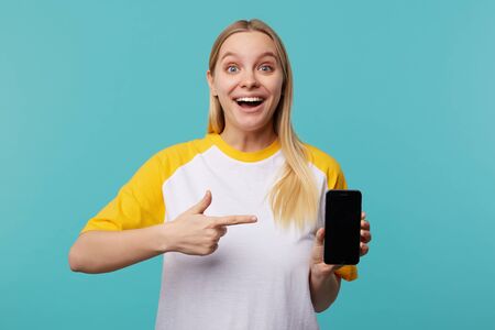 Open-eyed young pretty long haired white-headed woman pointing joyfully on her smartphone with raised index finger and smiling widely at camera, posing over blue backgroundの写真素材