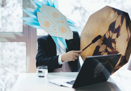 Asian businessman with tambourine in shamanic mask. Concept of attracting success, wishing good luck to business. Money increasing. Occult ritual, spiritism. Sitting at the table with laptop.の写真素材