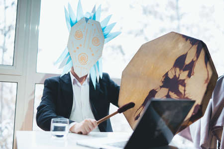 Asian businessman with tambourine in shamanic mask. Concept of attracting success, wishing good luck to business. Money increasing. Occult ritual, spiritism. Sitting at the table with laptop.の写真素材
