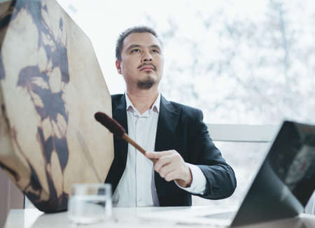 Asian businessman with shamanic tambourine. Concept of attracting success, wishing good luck to business. Money increasing. Occult ritual, spiritism. Sitting at the table with laptop.の写真素材