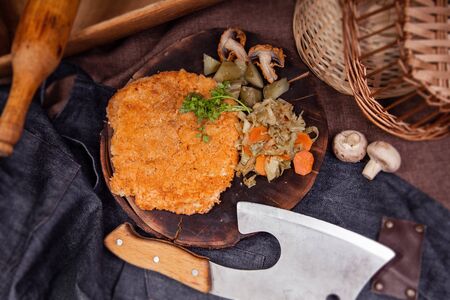 Breaded chop with mushrooms champignons and sauerkraut on a board with a knife on the background of an apron and rolling pinの写真素材