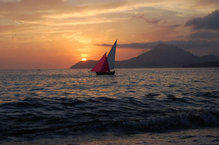 Photo of a sailboat sailing on the sea with stormy waves and stony shore. Ship on sunset with sky in purple pink orange colors and cloudsの写真素材