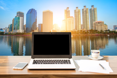 Mockup image of laptop with blank white screen on wooden table view outdoors of office building cityscape background.の写真素材