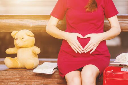 Asian woman pregnant red dress Heart-shaped handmade sitting on a bench with Teddy Bear and map with red suitcase at railway station travel.の写真素材
