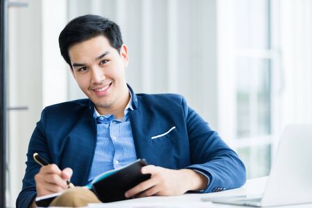 Happy mood a cheerful of asian young businessman working with making notes in a notebook and laptop computer  In the office room background.の写真素材