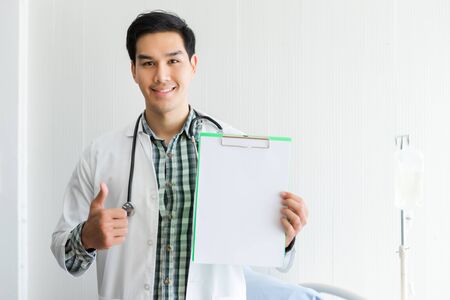 Asian Young man doctor therapeutic advising with positive emotions hold empty clipboard with showing thumbs up and bed in hospital background,copy spaceの写真素材