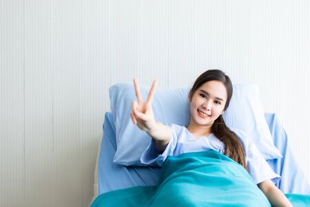 Asian young female patient smiley face lifts two fingers up fighting with illness on bed in the room hospital background.の写真素材