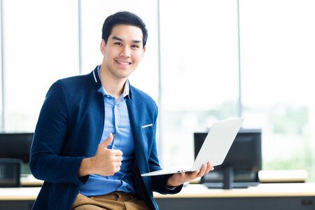 Happy successful mood asian young businessman showing thumbs up working with laptop computer sit on the table In the office room background.の写真素材