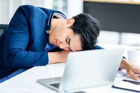 Asian young businessman worked late and fell asleep on laptop computer In the office room background.の写真素材