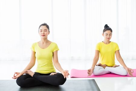 Two young Asian women workout practicing yoga in yellow dress or pose with a trainer and practice meditation wellness lifestyle and health fitness concept in a gym.の写真素材