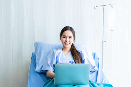 Asian young female patient with a smile working with laptop computer on bed in the room hospital background,concept of working business in a sick timeの写真素材