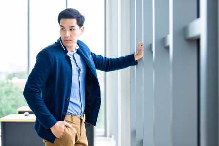 Portrait of a cheerful mature asian young businessman wear a business suit of man in blue jacket and blue shirt looking at the window In the office room background.の写真素材