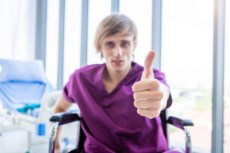a man patient sit in a wheelchair Abstract blur with focus show showing thumbs up with smiley face very good symptom in hospital background.の写真素材