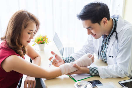 Asian Young man doctor of checking splint the arm and syringe to the arm of female patient hand due to with her arm broken for better healing sit in a wheelchair In the room hospital background.の写真素材