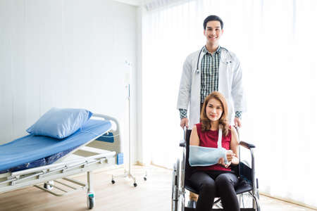 Asian Young man doctor of checking splint the arm of female patient hand due to with her arm broken for better healing with a smile sit in a wheelchair In the room hospital background.の写真素材