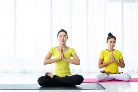 Two young Asian women workout practicing yoga in yellow dress or pose with a trainer and practice meditation wellness lifestyle and health fitness concept in a gym.の写真素材