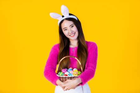 Easter holiday concept, Happy Asian Young woman wearing bunny ears hand holding a basket with colorful Easter eggs In isolated on Yellow blank copy space studio background.の写真素材