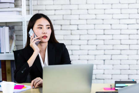Happy of successful asian young businesswoman hold pick up smartphone working with laptop computer in on White wooden table in white wall office backgroundの写真素材