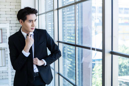 Portrait of a cheerful mature asian young businessman wear a business suit of man in blue jacket and blue shirt looking at the window In the office room background.の写真素材