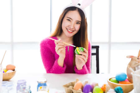 Easter holiday concept, Happy Asian Young woman wearing bunny ears hand painted eggs for Easter with colorful Easter eggs In the white room backgroundの写真素材