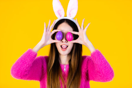 Easter holiday concept, Happy smile Asian Young woman wearing bunny ears and holding up a colorful Easter egg in front of her eye Skewers In isolated on Yellow blank copy space studio background.の写真素材