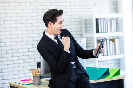 Excited overjoyed successful feeling winner screaming young Asian businessman holding working with a table In the office room background,Business successの写真素材