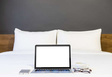 Laptop computer with blank white screen with smartphone,coffee cup and spectacles on white bed decoration in hotel bedroom interior background,Work and business in leisure with travel in the holiday.の写真素材
