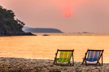 Beautiful early sunset over two deck chairs on the sandy tropical beach and Wave of the sea on the...