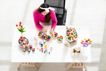 Easter holiday concept,Happy Asian Young woman wearing bunny ears hand painted eggs for Easter with colorful Easter eggs,basket Easter eggs,paint palette and brushes on white wooden table backgroundの写真素材