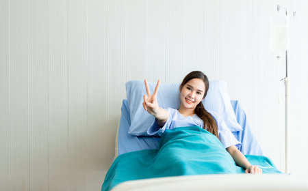 Asian young female patient smiley face lifts two fingers up fighting with illness on bed in the room hospital background.の写真素材
