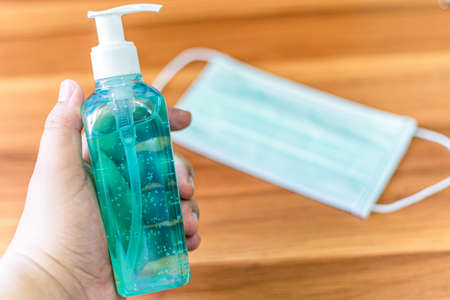 Hand holding of blue alcohol hand sanitizer Abstract blur of Protective disposable medical mask on wooden In the room at home background,Concept of Prevention of the spread of COVID-19 virus infectionの写真素材