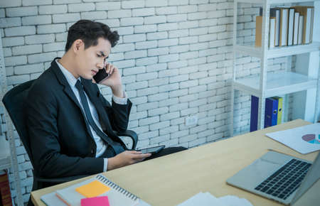 Stressed businessman hold pick up smartphone worked with laptop computer and having a headache after business losses In the office room background.の写真素材