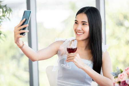 Selfie using a smartphone of Happy of smiling Asian young female sitting at a table food holding with wine glasses at in the restaurant backgroundの写真素材