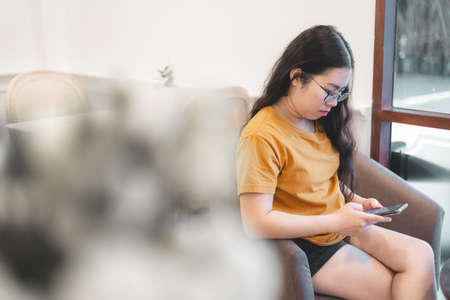 asian young woman in sitting on the chair and writing message on smartphone with window in the cafe or homeの写真素材