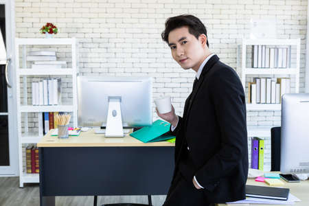Asian young businessman see a successful business plan holding a coffee cup,laptop computer on wooden table background in office,business expressed confidence embolden.の写真素材