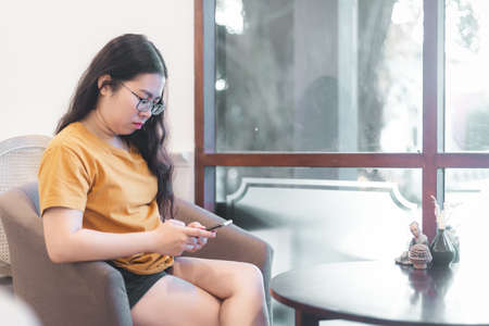 asian young woman in sitting on the chair and writing message on smartphone with window in the cafe or homeの写真素材