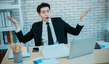asian young handsome businessman have surprised see a the laptop computer and document business plan on wooden table after business losses and unable to meet client needs In the office room backgroundの写真素材