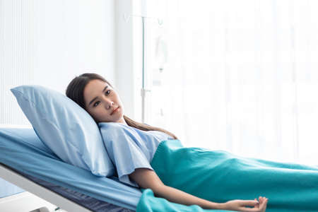 One asian young female patient With anxiety face lying and saline are lying on bed in the room hospital background.の写真素材