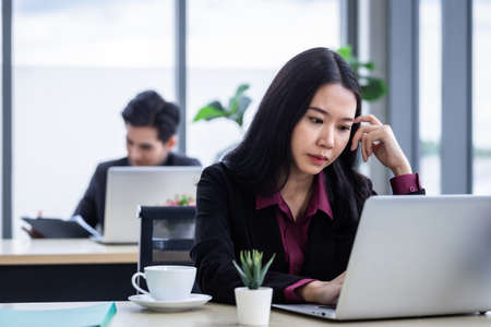 Upset thoughtful Young Asian business woman stress in the workplace working with laptop computer of Office staff are not happy in with working colleagues behind in the officeの写真素材