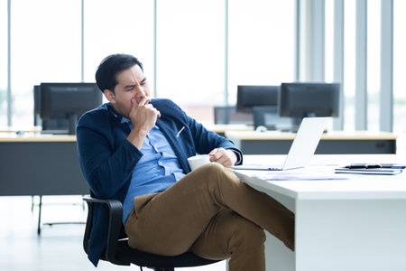 Asian young businessman bored yawning in front worked late and tired fell asleep or having stressful time on laptop computer In the office room background.の写真素材
