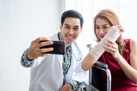 Selfie of smiling man doctor with a female patient wear arm splint for better healing sit in a wheelchair using a smartphone In the room hospital background.の写真素材